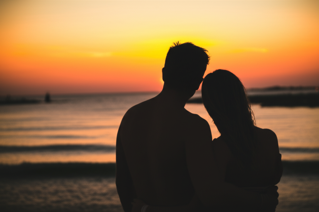 Um casal abraçado, de costas, observando o pôr do sol dourado na praia da Porto da Barra, com o mar calmo à frente.
