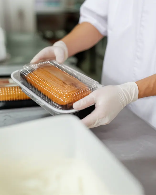 Confeiteiro com luvas, embalando cuidadosamente um bolo caseirinho em uma caixa transparente, foco na precisão