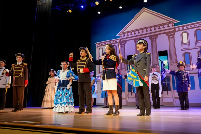 Alunos encenando a Proclamação da República Alunos vestidos com trajes de época encenam a Proclamação da República em um palco escolar, com cenário histórico.